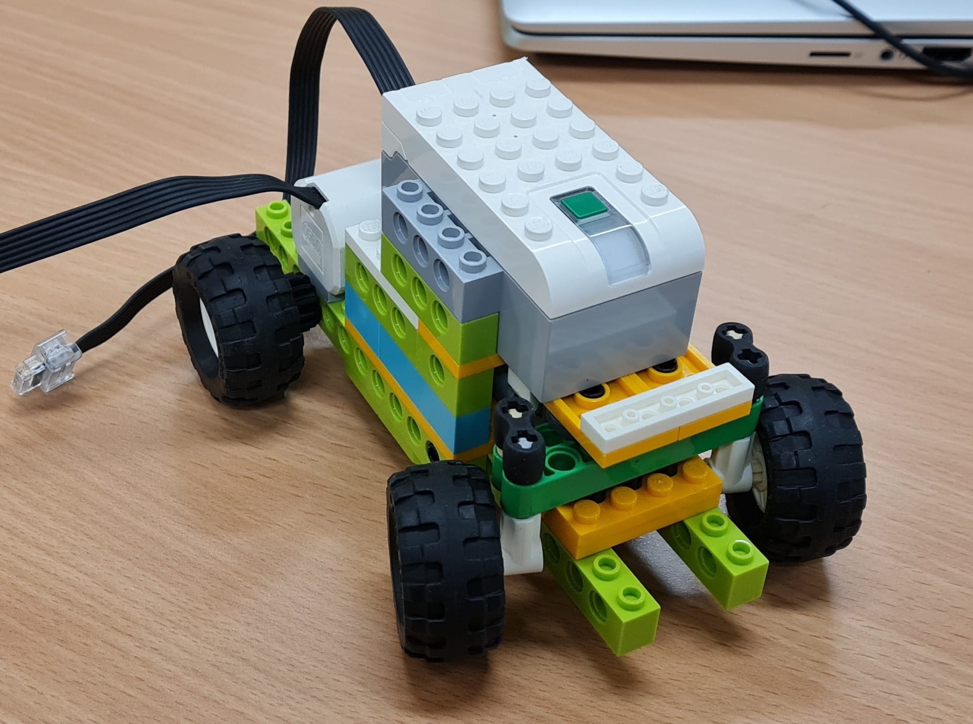 A four-wheeled LEGO robot built with green, yellow, and white bricks, connected to a black cable on a wooden desk.
